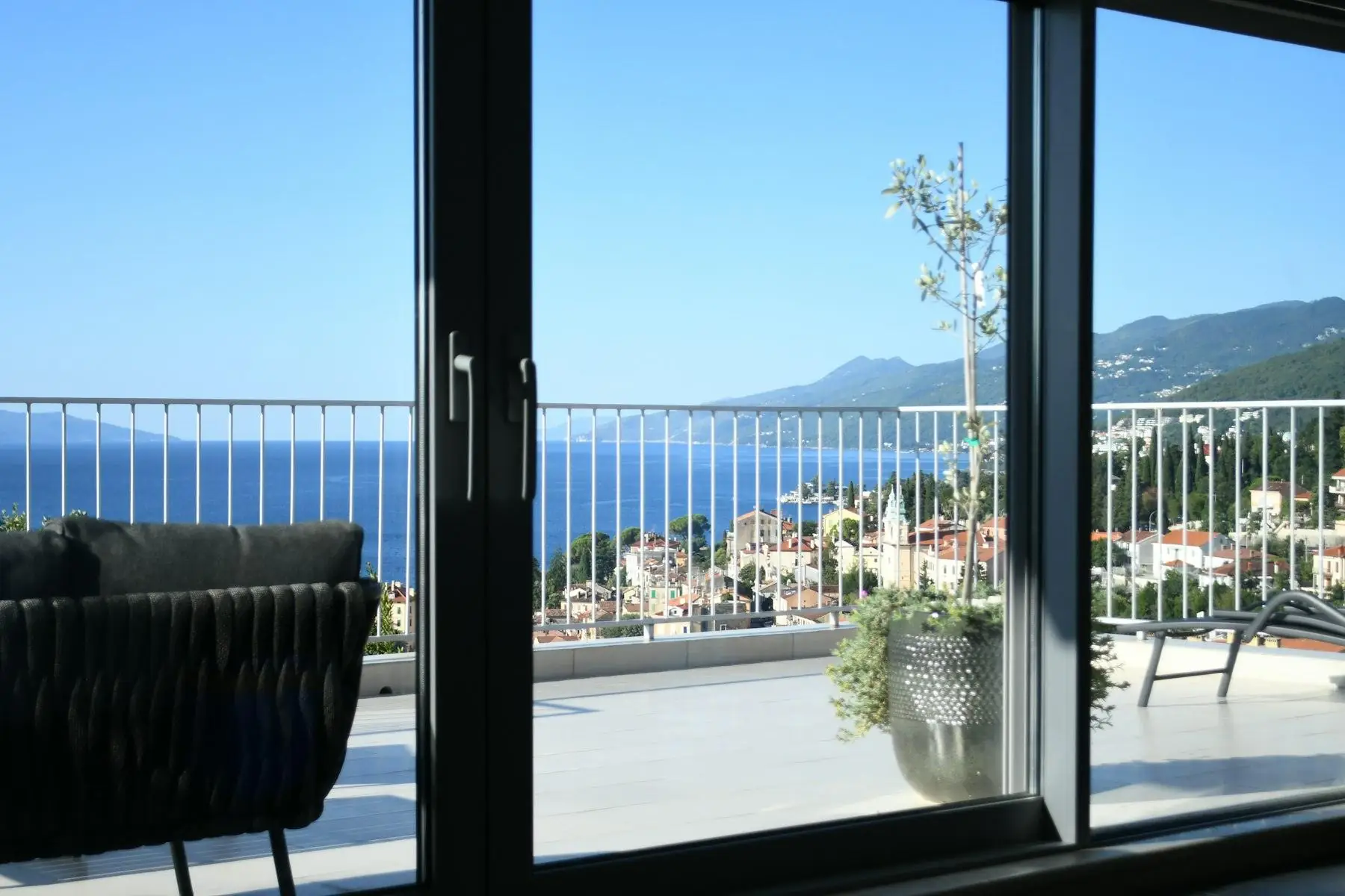 Vista dall’interno attraverso una portafinestra scorrevole dal telaio scuro: una terrazza con un ulivo in vaso, il mare e un paese sulla costa