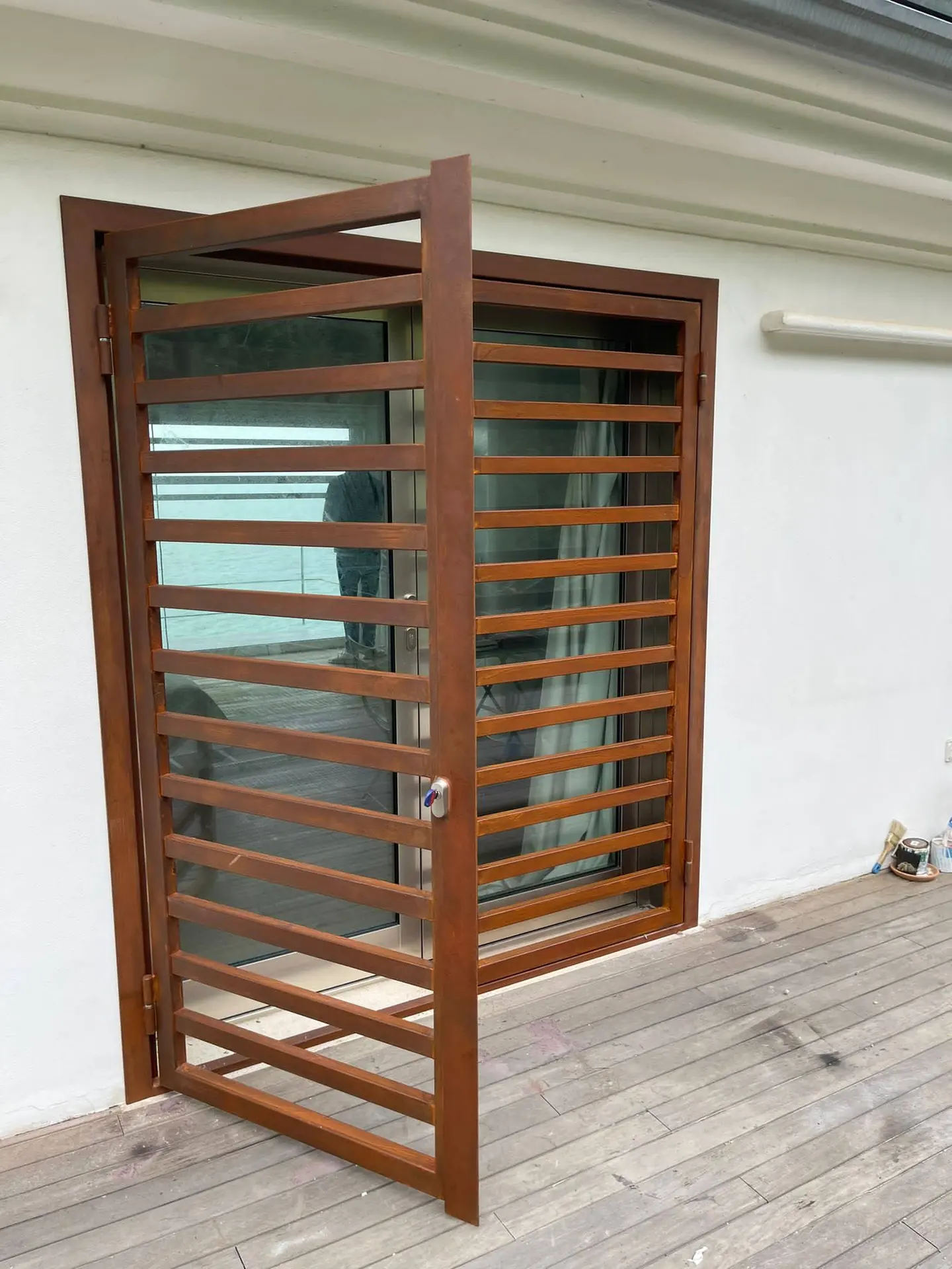 Grata apribile color ruggine aperta davanti a una portafinestra, su un muro bianco e un pavimento in legno