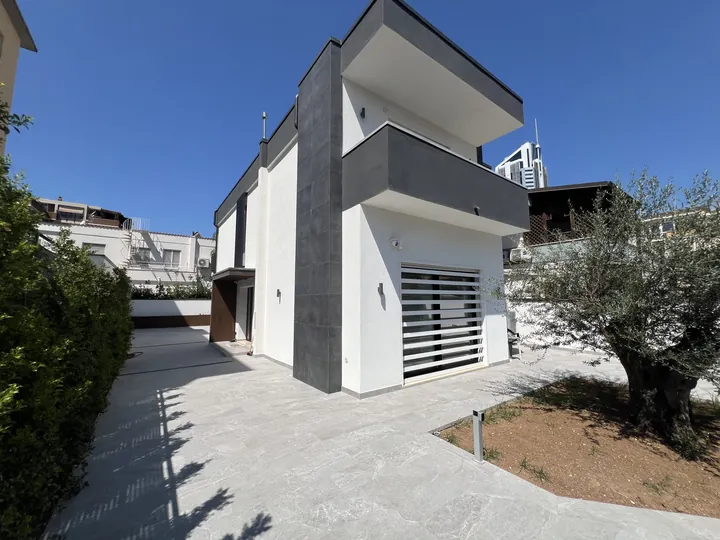 Casa moderna con loggia scura e grata bianca a lamelle al piano terra, tra un ulivo e la siepe